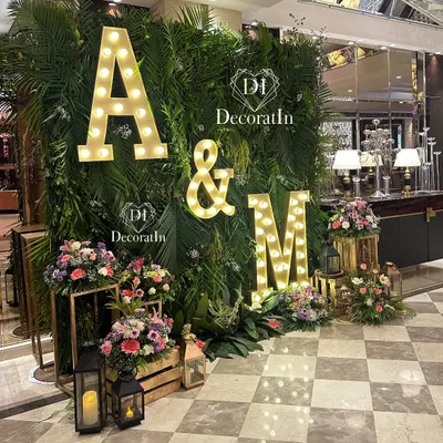 A stunning engagement photo wall featuring a lush backdrop of tropical greenery. The couple's initials, "A & M," in large marquee lights, create a personalized and glowing focal point for pictures.