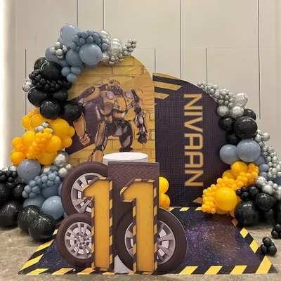 An action-packed Transformers theme for an 11th birthday, featuring a Bumblebee backdrop and a large, custom-made number '11' with a tire-track design. The scene is framed by a balloon arch in signature yellow, black, and silver, ready for an Autobot celebration.