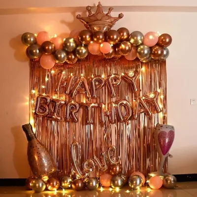 A very popular birthday decoration in a rose gold and gold theme. This setup includes a shiny foil curtain, 'Happy Birthday' letters, a balloon arch topped with a crown, and other fun balloons like a champagne bottle and a 'Love' sign.
