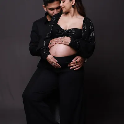 An intimate and stylish studio portrait. The all-black outfits create a chic, modern look, allowing the focus to remain entirely on the couple's tender embrace and the beautiful baby bump.