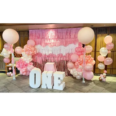 This is the full view of our fringe wall installation for a first birthday. The setup includes large 'ONE' marquee letters, two large balloon columns, and the hand-built pink, white, and rose gold fringe backdrop, creating a beautiful focal point.