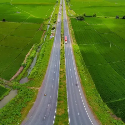A perfectly symmetrical drone shot of a highway cutting through vast, vibrant green paddy fields in rural Karnataka. The aerial perspective reveals the beautiful patterns of the landscape.