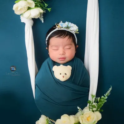 A sweet baby girl sleeps peacefully in a floral swing adorned with white peonies. The deep teal background makes the white flowers and the baby's teddy bear wrap pop.