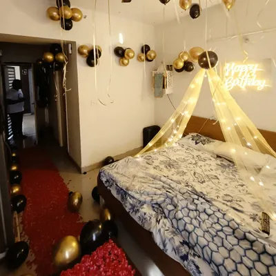 A romantic birthday room surprise with a black and gold theme. The setup includes a bed canopy with fairy lights, a neon sign, a red rose petal pathway, and balloons on the ceiling.