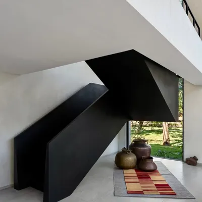 This staircase in the Mango Orchard House is designed as a bold, black, folded-metal sculpture. It creates a powerful visual statement against the light-colored walls, with a large window at the base framing a view of the garden.