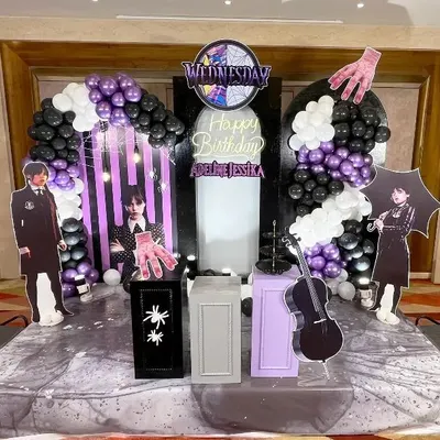 An elaborate 'Wednesday' themed birthday decoration featuring multiple character cutouts, a custom neon sign, and themed props. The black, white, and purple balloon decor creates a striking visual centerpiece for the party.