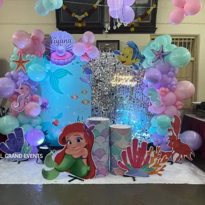 A complete 'Under the Sea' birthday party setup for Viyana. The scene is set with a silver shimmer wall, character cutouts of Ariel and Flounder, and custom props like coral and a treasure shell.