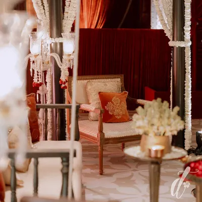 A close-up of a seating area from a royal-themed reception. The decor features elegant candelabras draped with jasmine, set against a backdrop of rich, red velvet curtains.
