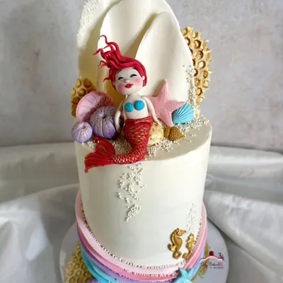 This mermaid cake is a more artistic take on the theme, featuring a hand-sculpted mermaid with vibrant red hair resting against a large, white chocolate seashell. Edible sand, seahorses, and coral details complete the underwater scene.
