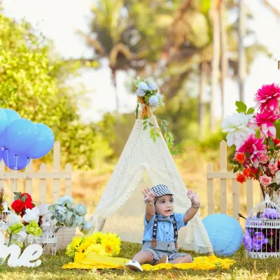 A baby boy enjoys an outdoor photoshoot in a beautiful, sunny field. The setup includes a lace teepee, colorful flowers, and balloons, creating a perfect picnic-like atmosphere for his first birthday.