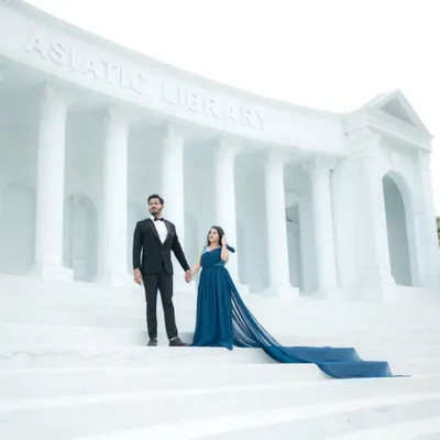 A majestic wide shot of a couple standing on the steps of an Asiatic Library replica set. The groom in a sharp tuxedo and the bride in a gown with a long train create a truly grand and aspirational pre-wedding portrait.