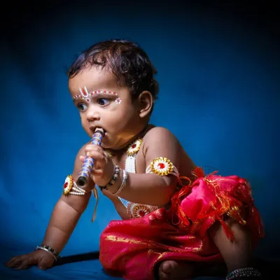A full-body shot from a 'Little Krishna' photoshoot. Here, the baby is captured in a classic pose with his flute against a dramatic blue background, highlighting the traditional red dhoti.