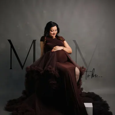 A classic and elegant studio portrait. The mother-to-be is seated in a dramatic, ruffled brown gown, looking down at her bump with a gentle smile, embodying the grace of this special time.