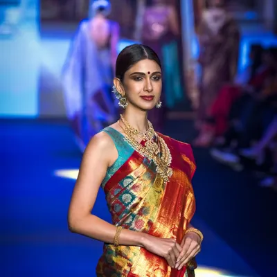 A classic runway shot from Bangalore Times Fashion Week, capturing the model's confident walk. The focus is on the traditional silk saree, showcasing its vibrant colors and elegant drape under the stage lights.