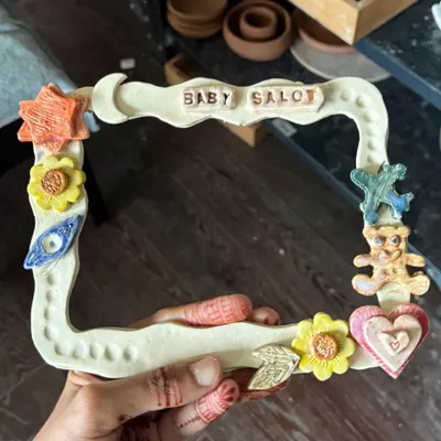 A close-up of a child's creation: a colorful, hand-built ceramic photo frame decorated with stars, flowers, and a teddy bear.
