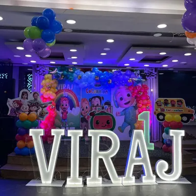 A vibrant Cocomelon stage setup for Viraj's first birthday. The decor features a large character backdrop, rainbow balloon columns, and light-up marquee letters for the name.