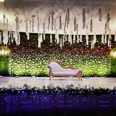 This outdoor reception stage features a massive wall of red and white flowers, creating a dramatic and romantic backdrop. I used hanging white floral strings from the ceiling and spotlights on the greenery pillars to add depth and texture to the design.