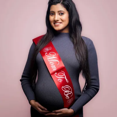 A classic studio maternity portrait. The simple pink background and the "Mom To Be" sash keep the focus on the expectant mother's happy expression.