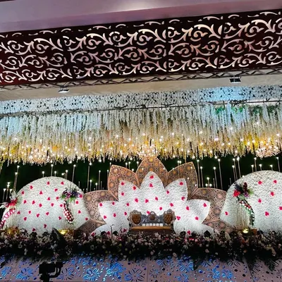 A grand reception stage featuring a lotus-inspired design made with white and red flowers. The cascading floral ceiling and intricate lighting create a truly breathtaking effect for a wedding reception.