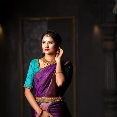 A dramatic portrait featuring a beautiful purple Kanchipuram silk saree. The lighting is designed to sculpt the subject and make the traditional jewelry and fabric shine.