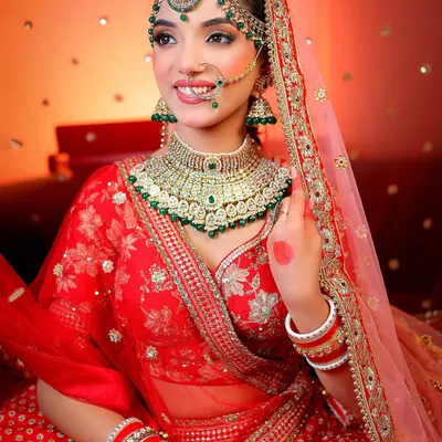 A smiling bridal portrait with a warm, celebratory background. The focus is on the intricate green and gold jewelry and the bride's happy expression.