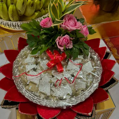 A classic sweet platter featuring Kaju Katli, presented on a silver tray with a red velvet lotus base. The arrangement is topped with fresh pink roses for an elegant touch.