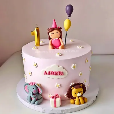 A pastel pink first birthday cake for little Aadhira. The design features cute fondant animals, a personalized name plaque, and a number '1' candle, making it a sweet and memorable centerpiece.