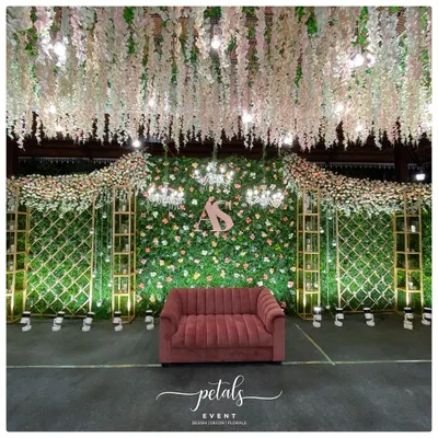 A luxurious reception stage featuring a lush green wall adorned with pastel flowers and the couple's monogram. The plush velvet sofa and cascading floral ceiling create a look of pure opulence and romance.