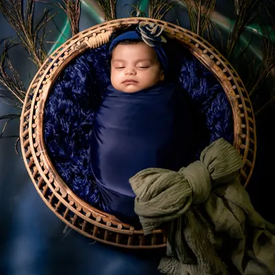 A peaceful newborn swaddled in royal blue, nestled in a rustic basket surrounded by peacock feathers. This is a classic example of a creative newborn setup.