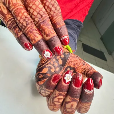 A stunning set of bridal nails designed to perfection. The deep red polish is accented with 3D white flowers and sparkling stones, creating a beautiful contrast with the intricate mehndi.