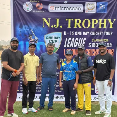 The opening ceremony of the N.J. Trophy U-15 cricket tour, with organizers and a young player posing in front of the official banner.