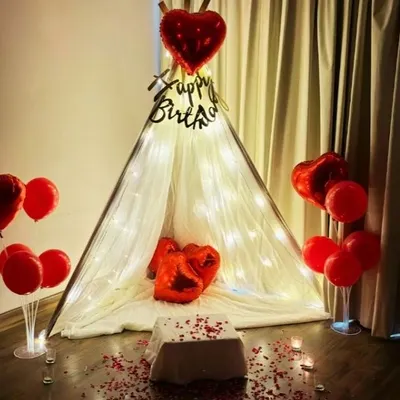 A romantic and intimate birthday surprise created right in the bedroom. This cozy setup features a beautiful teepee tent draped in sheer fabric and fairy lights, with red heart-shaped balloons and scattered rose petals to create a special, personal celebration.