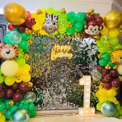 A jungle-themed first birthday party backdrop. This setup features a shimmering sequin wall, a 'Happy Birthday' neon sign, a light-up number '1', and a balloon arch with cute animal faces.