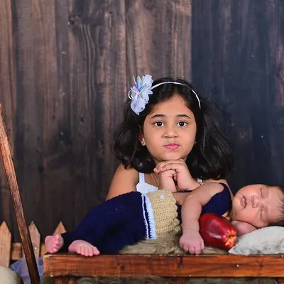 A big sister proudly watches over her sleeping newborn brother. This rustic studio setup with a wooden backdrop adds a warm, timeless feel to this sweet sibling portrait.