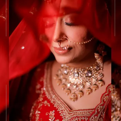 A serene and artistic bridal portrait. The bride's face is partially veiled by her red dupatta, drawing attention to her beautiful nose ring, intricate necklace, and peaceful expression.