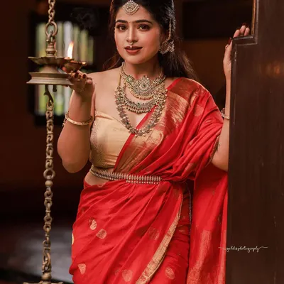 A classic portrait of a model in a red saree and gold blouse, holding a traditional lamp. The warm lighting and heritage setting create an atmosphere of timeless Tulunadu elegance.