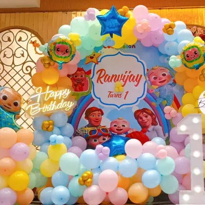 A vibrant and colorful Cocomelon theme for a first birthday. This setup features a custom character backdrop, a multi-colored balloon arch, and a large light-up number '1'.