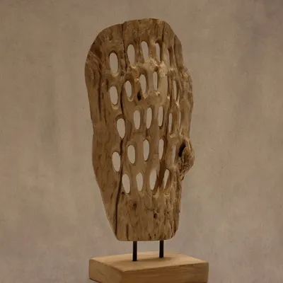 This handcrafted wood artifact features a unique perforated design, playing with light and shadow. Mounted on a simple teak base, it stands as a distinctive accent for any interior.