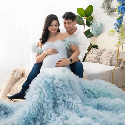 An intimate moment shared between the couple. The way they look at each other while cradling the bump speaks volumes about their shared love and excitement. The soft blue gown and light-filled setting create a serene atmosphere.
