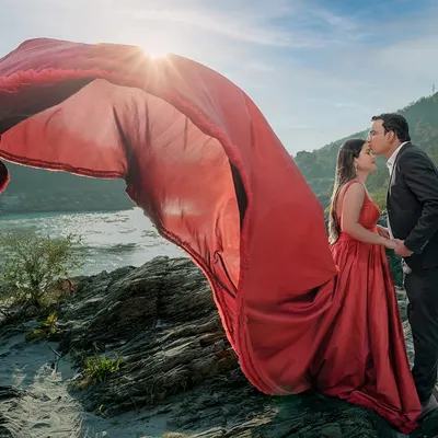 With the sun setting over the river, her red gown catches the wind, creating a moment of pure cinematic magic. This is the kind of epic, dramatic pre-wedding shot that becomes the poster for your love story.