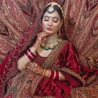 A regal bridal portrait in a deep red lehenga. The model is framed by the rich fabric, showcasing the intricate gold embroidery and a heavy choker set with green accents.