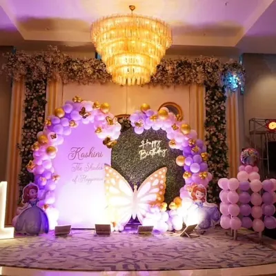A stunning stage setup in a large banquet hall for a butterfly-themed birthday. The grand chandelier complements the purple and gold balloon arch and custom backdrop, creating a luxurious feel.
