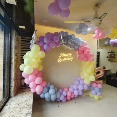 A beautiful pastel rainbow theme for a second birthday. This circular balloon arch uses soft shades of pink, yellow, purple, and blue, complemented by a 'Happy Birthday' neon sign.