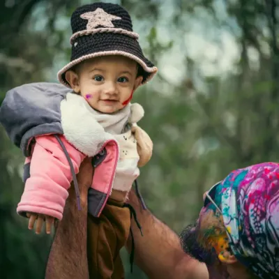 A father joyfully lifts his son into the air during his very first Holi celebration. The baby's smile and the colorful background perfectly capture the happiness of this family festival.