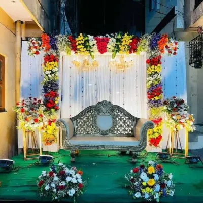 A vibrant and colorful stage decoration for an engagement, featuring a multi-colored floral arch, elegant chandeliers, and a classic gold-trimmed sofa.