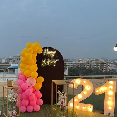 A chic 21st birthday decoration on a rooftop. This setup includes large illuminated marquee numbers, a "Happy Birthday" neon sign against a dark arch, and a bright pink and yellow balloon arrangement.