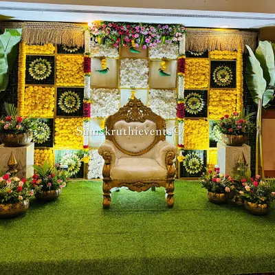 A classic and elegant backdrop for a housewarming ceremony. The design features a checkerboard pattern of yellow marigolds and white chrysanthemums, accented with woven leaf details and large banana plants.