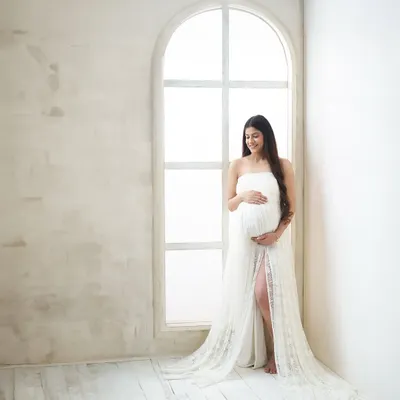 A full-length portrait using the beautiful natural light from an arched window. The bright, clean setting and elegant white lace gown create a serene and angelic atmosphere.