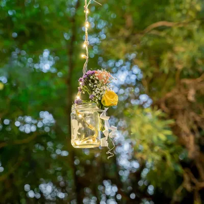 A simple mason jar filled with a small bouquet and fairy lights, hanging from a tree. This is a perfect example of how I use small, creative details to add a touch of rustic charm to an outdoor event.