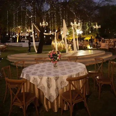 This photo captures the enchanting ambiance of an outdoor reception at night. A round table is set with a patterned linen, surrounded by wooden cross-back chairs, all under a canopy of twinkling fairy lights.